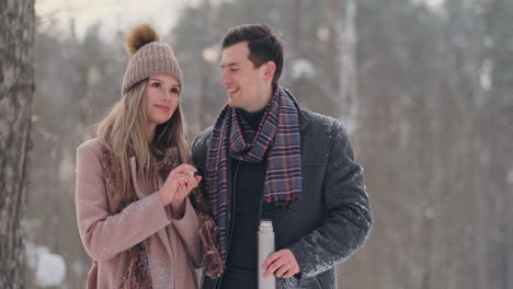 Couple-in-love-in-the-winter-forest-to-drink-tea-from-a-thermos.-Stylish-man-and-woman-in-a-coat-in-the-Park-in-winter-for-a-walk.